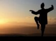 Chinese Taoist monk engaged in the practice of Qigong atop a tranquil mountain during sunset. The monk is standing in a classic martial arts pose, demonstrating the harmonious balance of energies - Jing, Qi, and Shen. His serene stance against the backdrop of the setting sun beautifully symbolizes the cultivation and refinement of energy within the Taoist practices of Neigong and Neidan, leading to improved health, unlocking of extrasensory abilities, and the path to enlightenment.